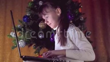 圣诞树下有笔记本电脑的女孩。 穿着白色汗衫的小女孩在新叶的圣诞树下玩笔记本电脑